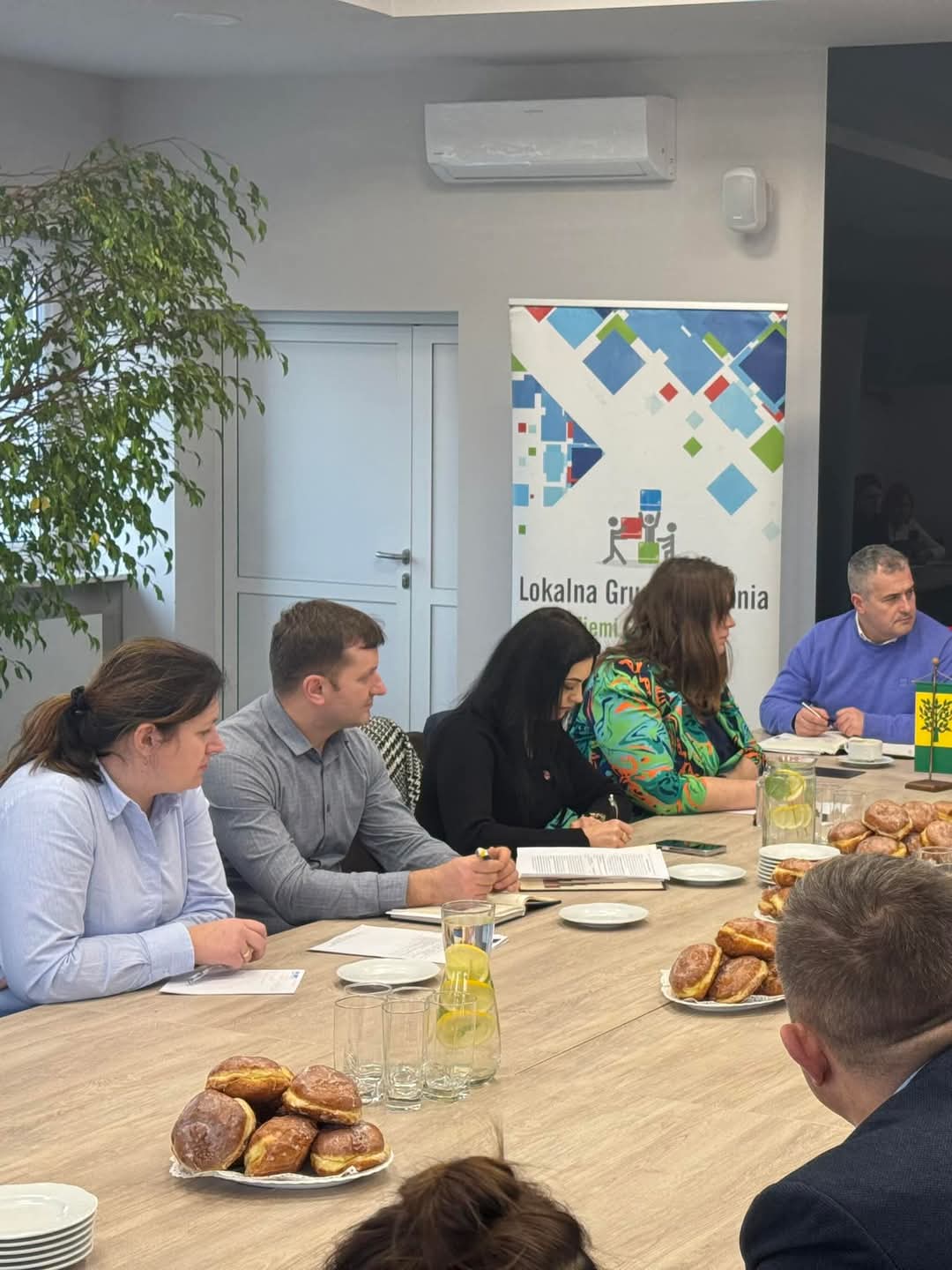 Oficjalne spotkanie w nowoczesnej sali konferencyjnej. Uczestnicy skupieni na dokumentach, atmosfera rzeczowa. W tle baner Lokalnej Grupy Działania Ziemi Siedleckiej.
