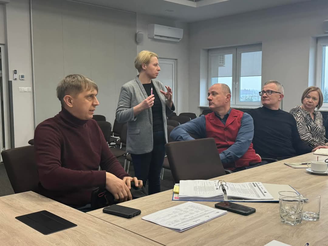 Oficjalne spotkanie w nowoczesnej sali konferencyjnej. Uczestnicy skupieni na dokumentach, atmosfera rzeczowa.